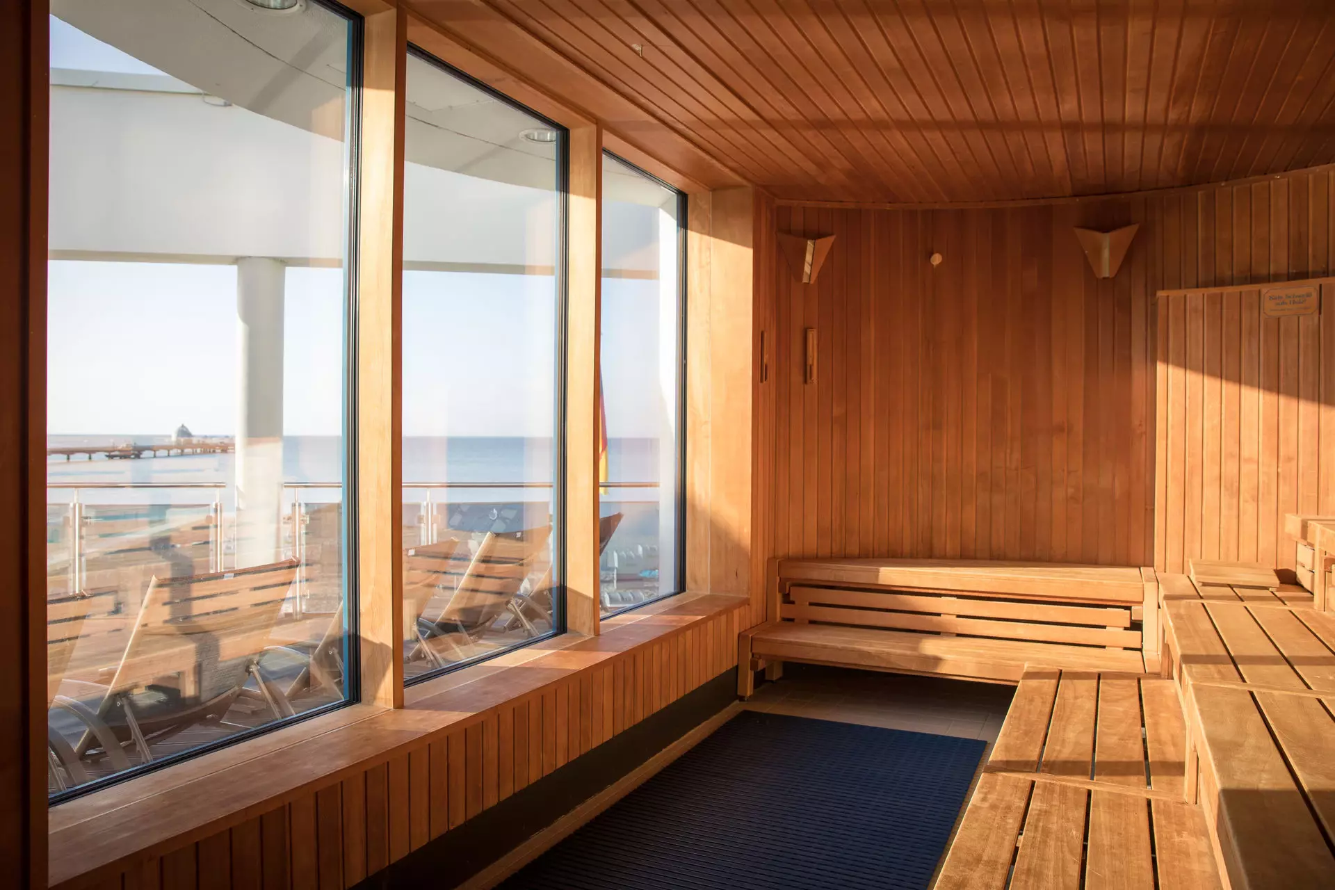 Ausblick aus einer hölzernen Sauna hinaus durch drei breite Panoramafenster auf den Sandstrand und das Wasser der Ostsee.