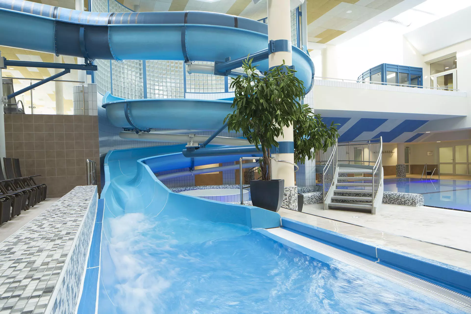 Endbereich einer blauen Wasserrutsche vor einer kleinen Treppe und einer grünen Pflanze im Innenraum eines Schwimmbads. Im Hintergrund ist die Röhrenrutsche zu mit zwei Kurven zu erkennen.