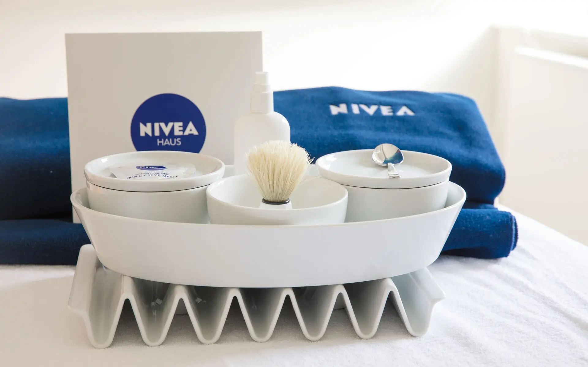 Vor zwei dunkelblauen, gefalteten Handtüchern mit NIVEA-Logo stehen in einer weißen, ovalen Schale drei weiße, runde Schüsselchen mit Deckel. Dazwischen ist eine weiße NIVEA-Tüte mit dunkelblauem NIVEA-Logo platziert.