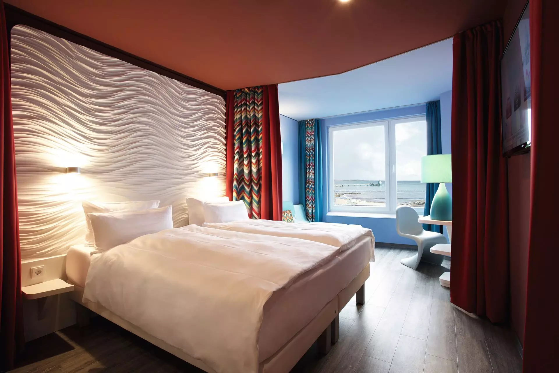 Ein mit weißer Bettwäsche bezogenes Doppelbett steht vor einer weißen Rückwand mit Struktur. Umrahmt von einer dunkelroten Wand und einem rot-gemusterten Vorhang steht das Bett in einem Raum mit rostroter Decke und dunklem Holzboden. Im Hintergrund ist ein von blauen Wänden umrahmtes Fenster mit Blick auf die Ostsee zu erkennen.